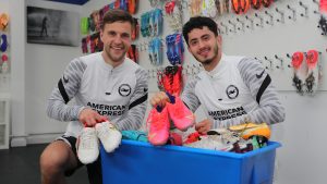 Steven Alzate and Joel Veltman crouched next to a box full of donated football boots, smiling and holding their own boots