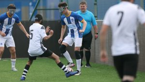 An Albion player stands with the ball at his feet while a Hereford player looks to block the ball. The referee looks on from the background.