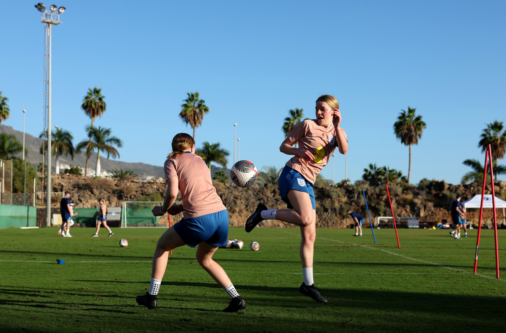 Faye, wearing pink England training kit, flicks the ball up on a grass pitch. It is sunny and there are palm trees in the background.