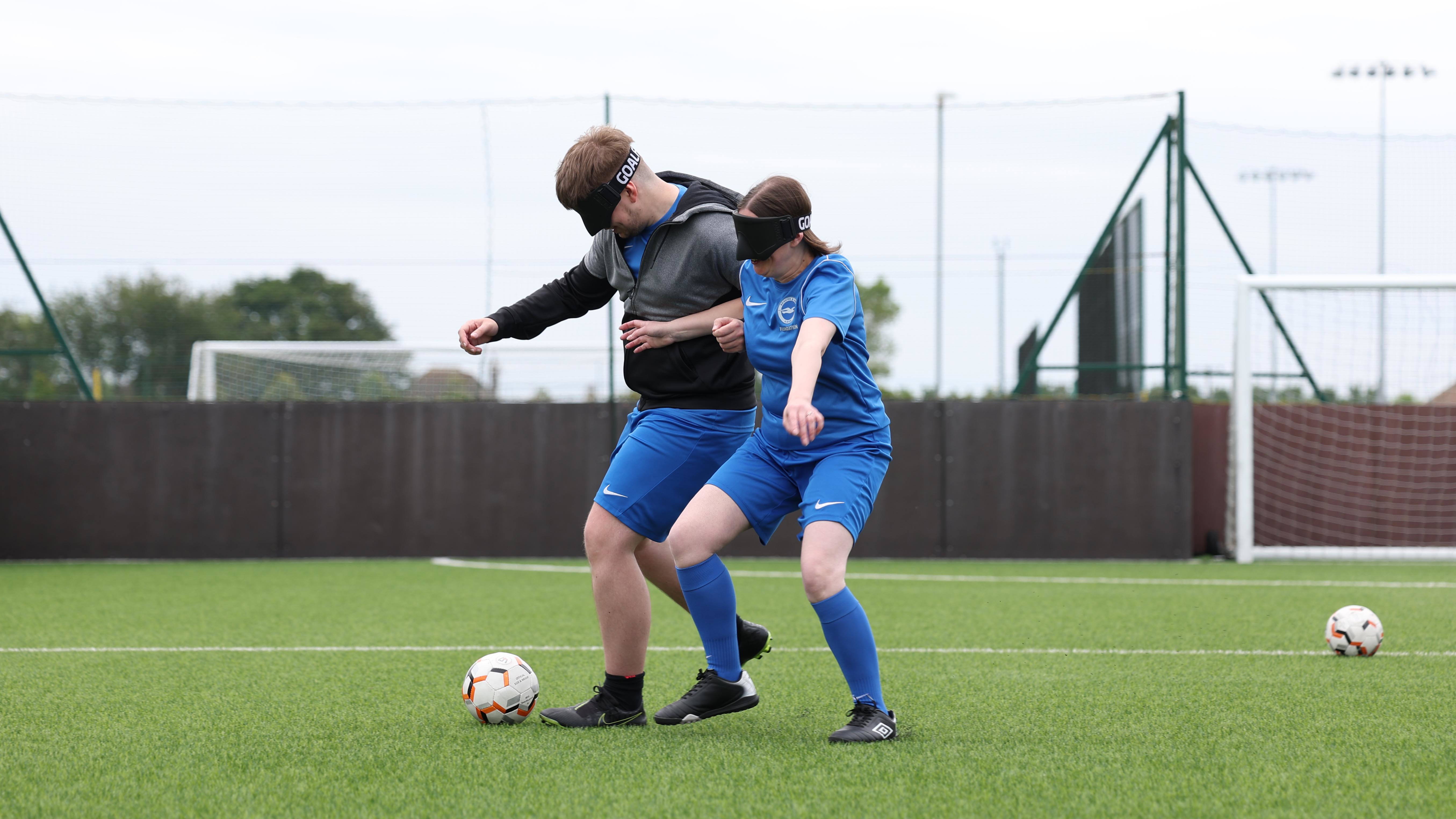 Alison and another player, Sam, jostle eachother to win the ball on a 3G football pitch. Both are wearing eyemasks.